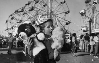 A young girl at a fairground holds a large teddy bear and excitedly eats cotton candy. Ferris wheels and crowds of people are visible in the background, capturing a festive atmosphere.