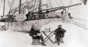 Two men bundled in heavy clothing sit on chairs at a small table set with drinks, on snow in front of a large, icebound ship; snowy landscape surrounds them.