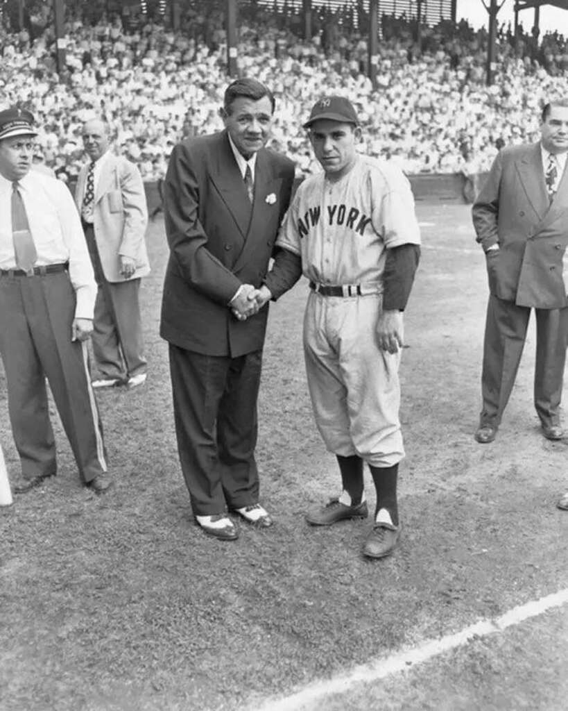 Two men shake hands on a baseball field, one in a suit and the other in a "New York" baseball uniform. A crowd fills the stands in the background, and several men stand nearby, some in suits and one in an umpire uniform.