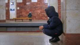 A person in a dark hooded coat squats near a pillar on a subway platform, while another person sits alone on a bench across the tracks. The station has marble walls and a quiet, empty atmosphere.