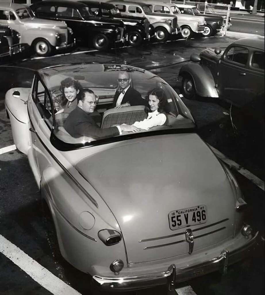 A vintage black and white photo shows four smiling people in a convertible car with the top down. The car has a California license plate. Other classic cars are parked in the background, suggesting a mid-20th-century setting.