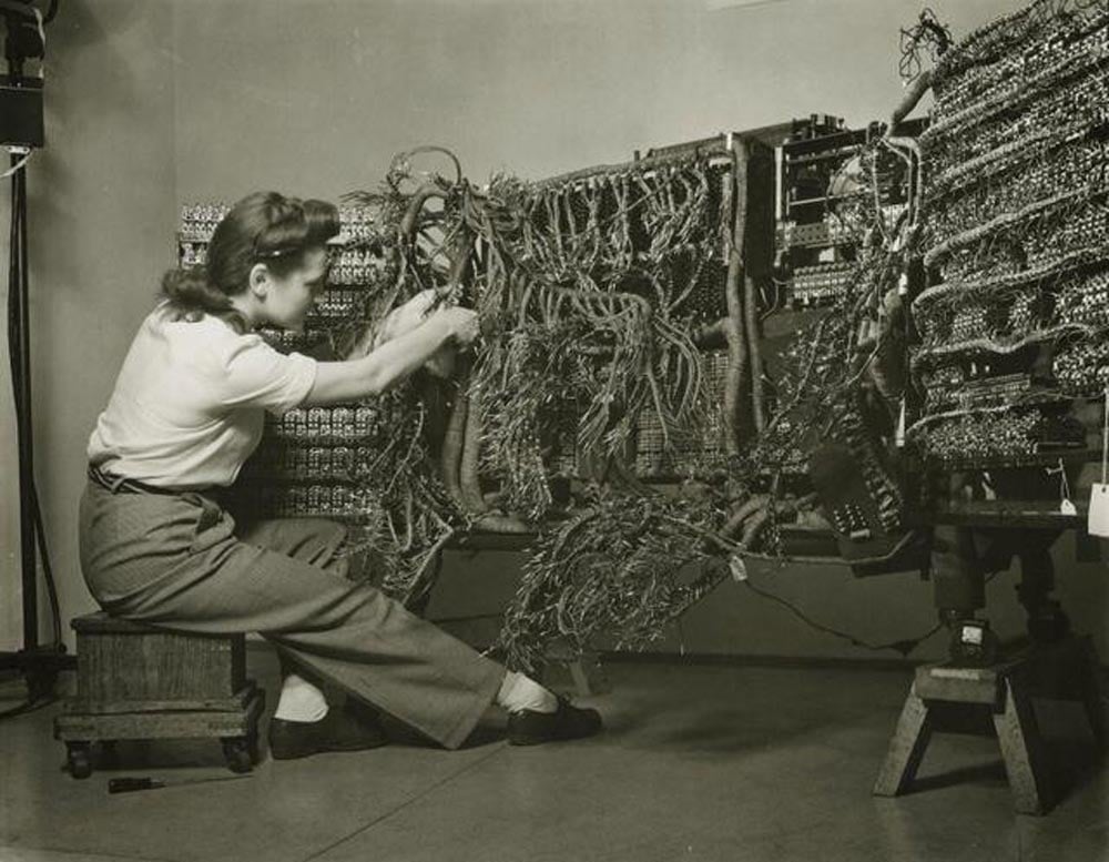 A woman is sitting on a small stool, working with a complex array of wires in a vintage computing machine. She is wearing a white shirt and trousers, surrounded by electronic components in a dimly lit room.