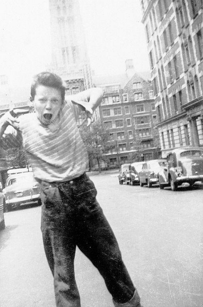 A boy energetically poses in the middle of a street, arms raised, with a playful expression. He wears a striped shirt and pants. Vintage cars line the street, and tall buildings frame the background on a sunny day.
