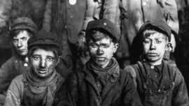 A black and white photo of three young boys with dirty faces, wearing worn-out clothes and caps, sitting closely together. Other children in similar attire are blurred in the background.