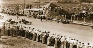 Historic racing scene with vintage race cars speeding down a track lined with spectators. A small grandstand and a crowd of people standing along the fence watch the action. The setting appears to be from an early 20th-century event.