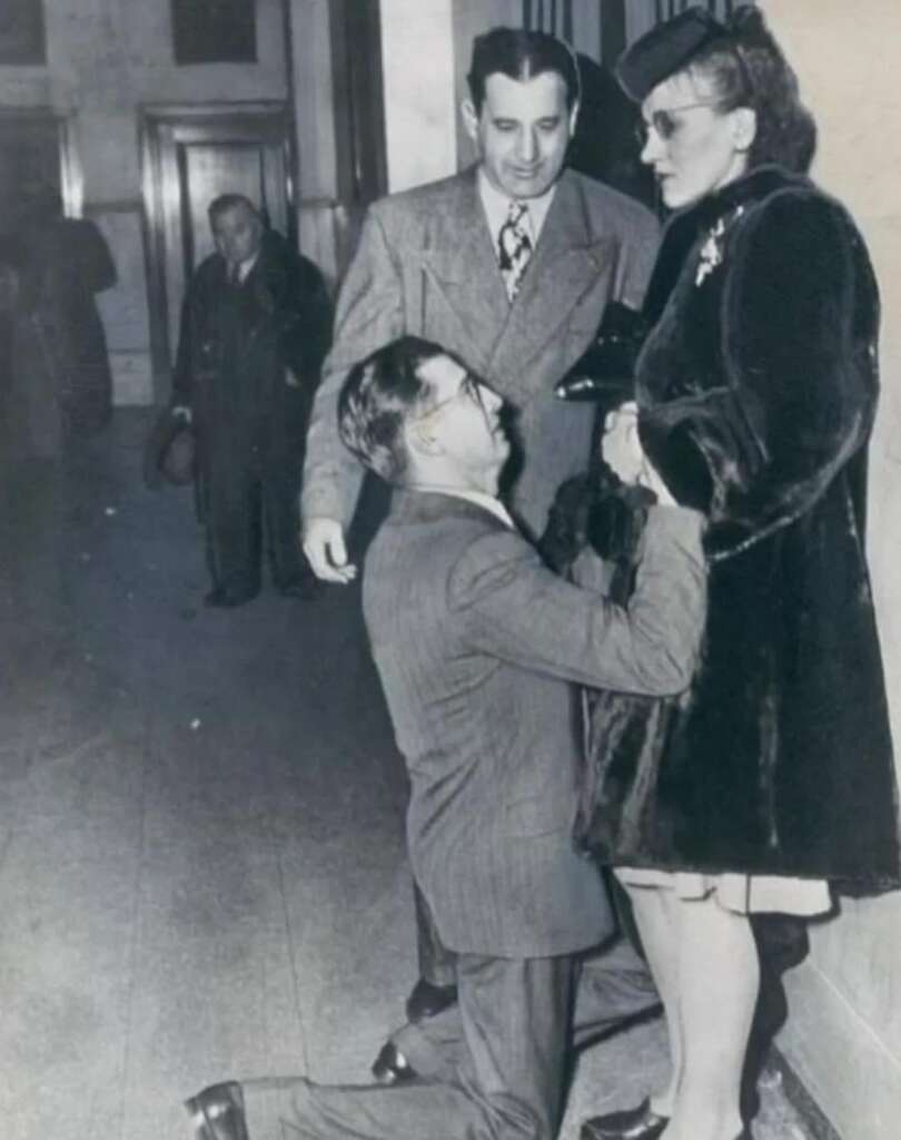 A man kneels on the floor holding the hand of a woman standing in a fur coat while another man stands beside them, smiling. They are in a hallway with another person in the background. The scene appears candid and from a bygone era.