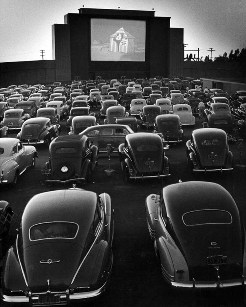 Vintage cars parked at a drive-in theater, watching a movie on a large screen in black and white. The film depicts a person wearing a cloak or robe. Tower lights are visible in the background.