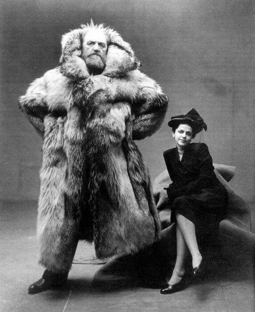 A man in a large, fur coat stands confidently with his hands on his hips, while a woman in a dark dress and hat sits beside him. The photo is in black and white, emphasizing the contrast and textures.