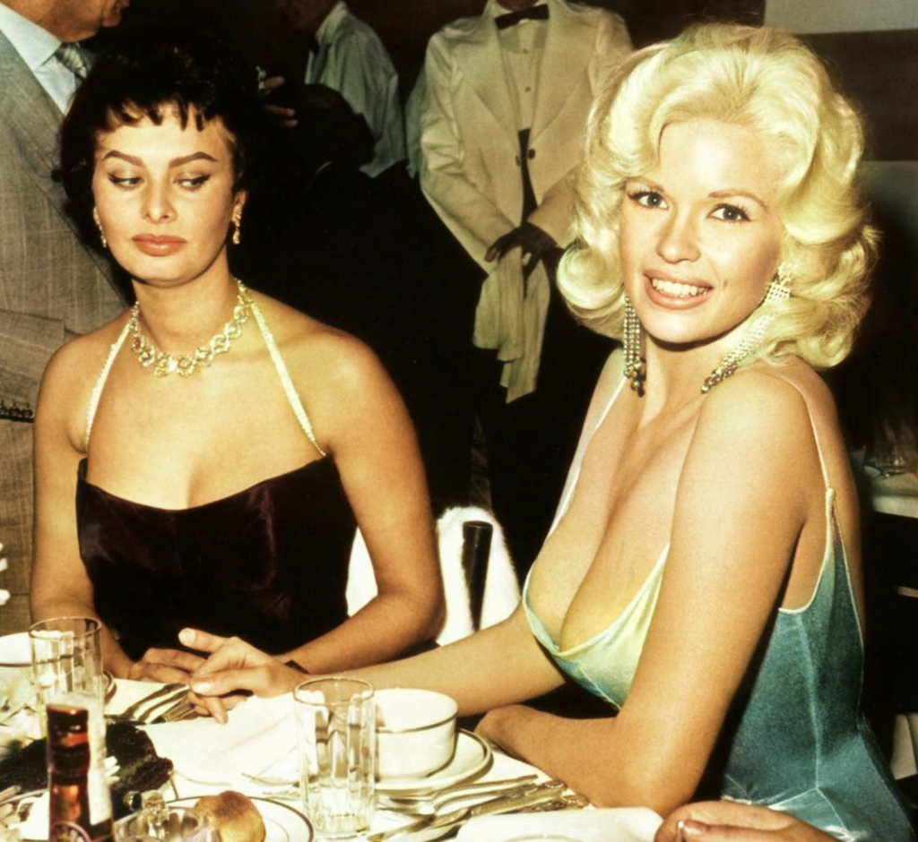 Two women in glamorous dresses sit at a formal dinner table. The woman on the left looks sideways with a reserved expression, while the blonde woman on the right smiles brightly at the camera.
