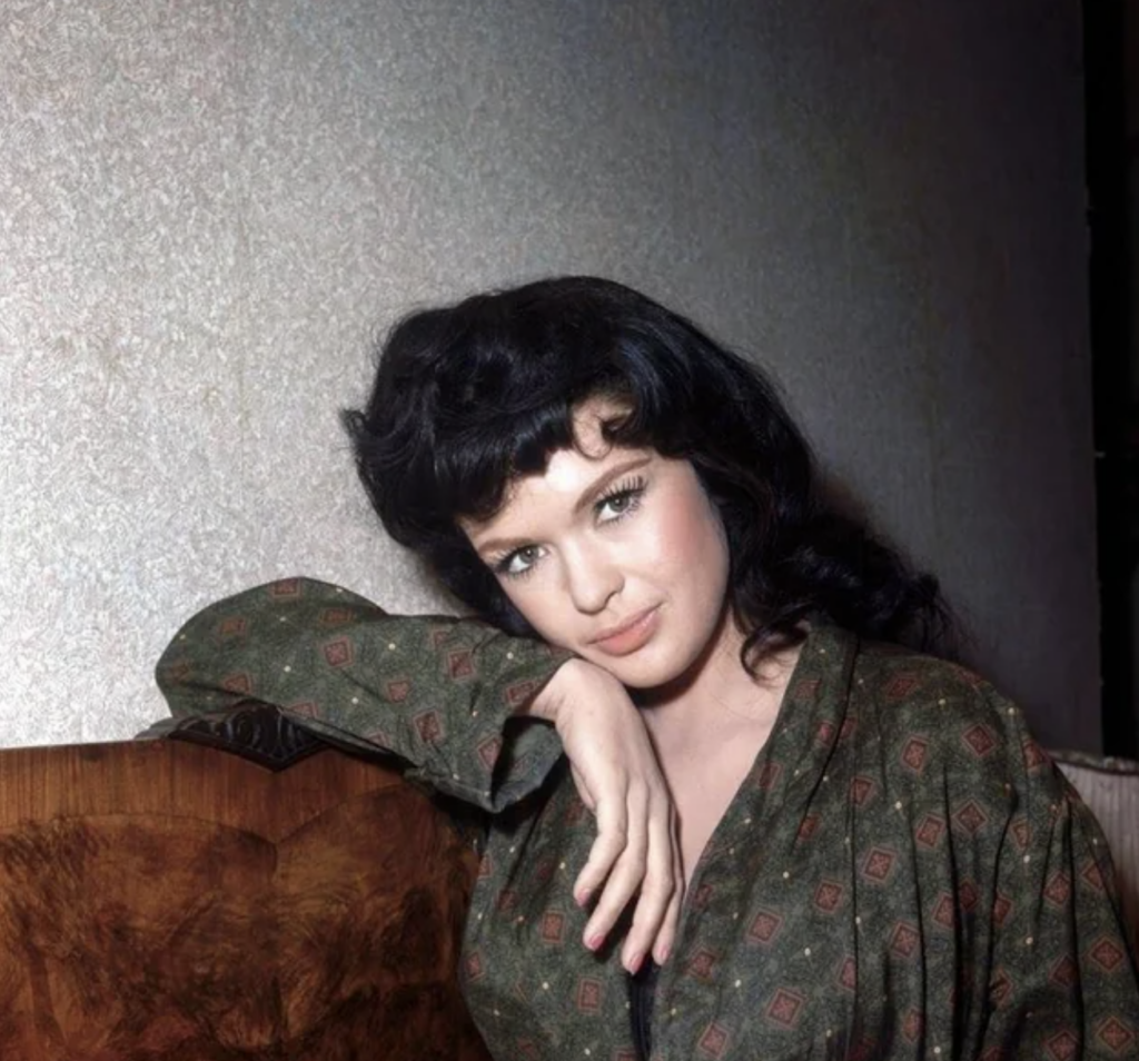 A woman with long dark hair and bangs leans her arm on the back of a wooden couch, looking at the camera. She wears a patterned green robe and sits against a light-colored textured wall.