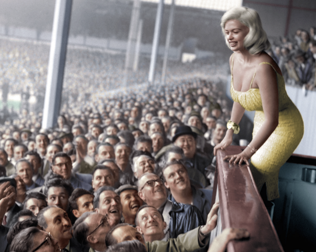 A woman in a yellow dress smiles and leans over a barrier to greet a large, cheering crowd of men at a stadium, many of whom are craning to get a closer look or take her hand.