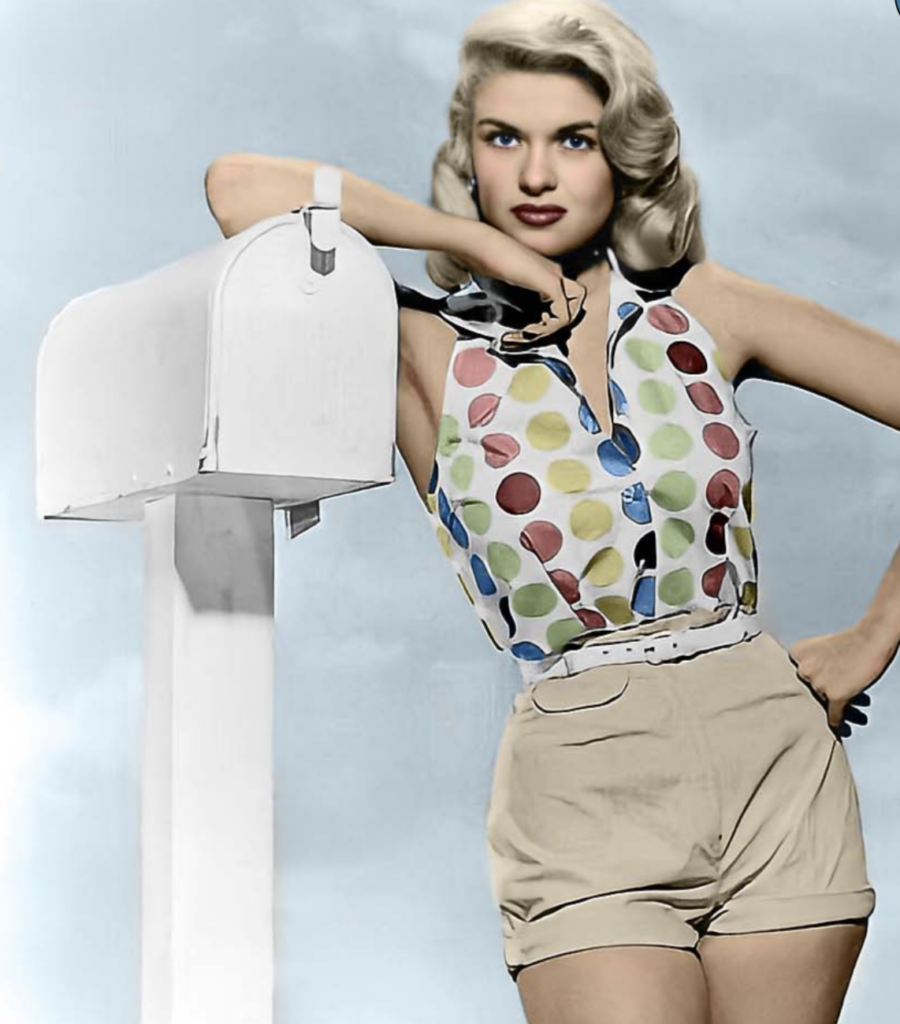 A woman with long blonde hair in a sleeveless, polka dot blouse and high-waisted shorts leans on a white mailbox, posing against a light blue sky.