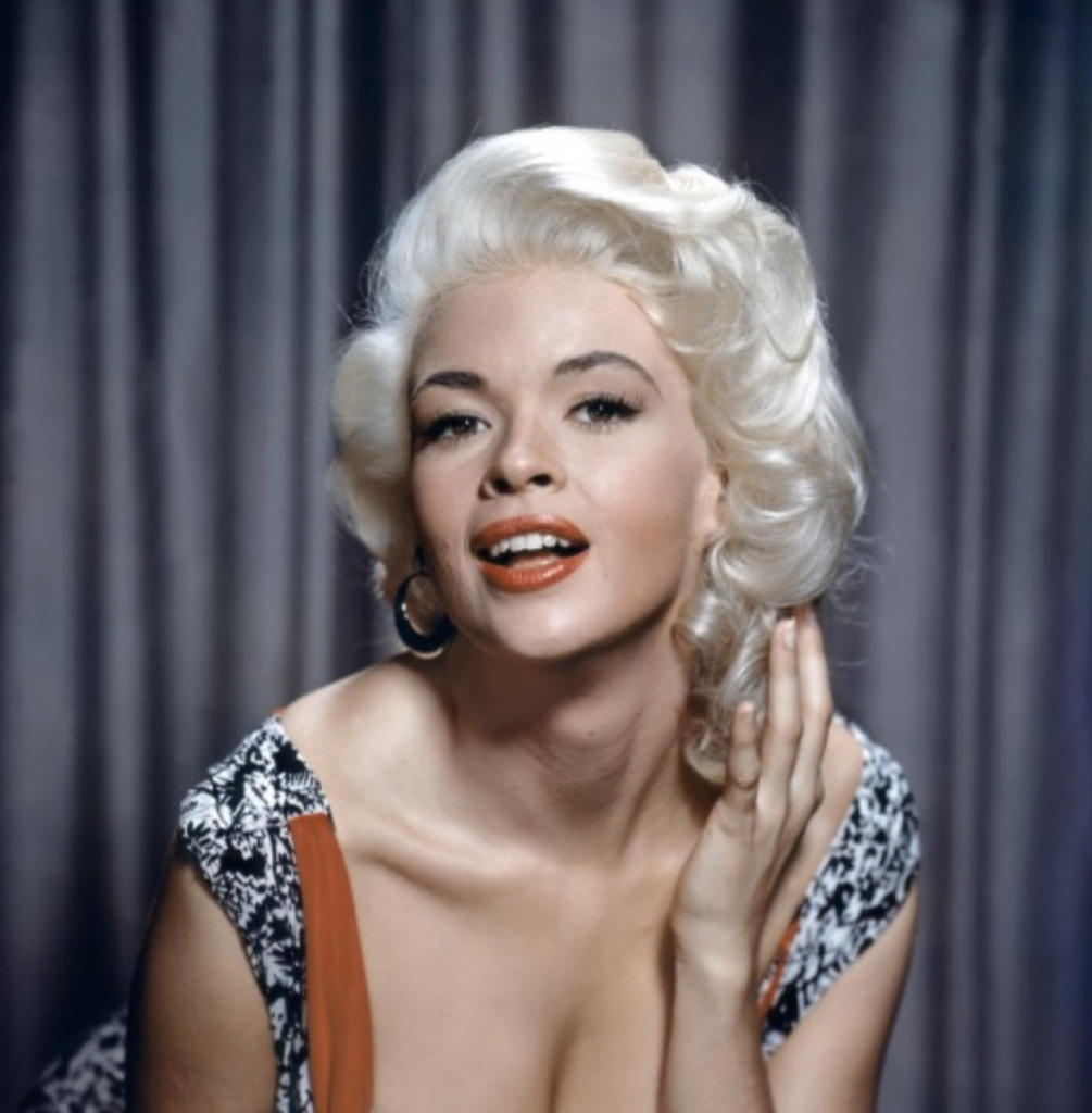 A woman with platinum blonde, wavy hair poses in front of a gray curtain, smiling with red lipstick. She is wearing a patterned dress with a low neckline and hoop earrings, with one hand touching her hair.