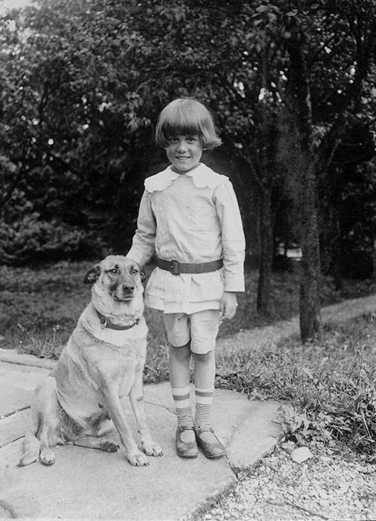 A child wearing a light-colored outfit stands beside a large dog on a stone path. Both look towards the camera. Trees and grass form the background, suggesting a park or garden setting.