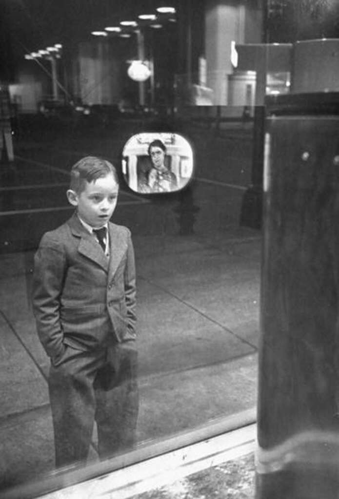 A young boy in a suit stands with his hands in pockets, looking through a window. A small, black-and-white television screen in the window displays an image of a woman. The street behind him is dimly lit with cars parked.