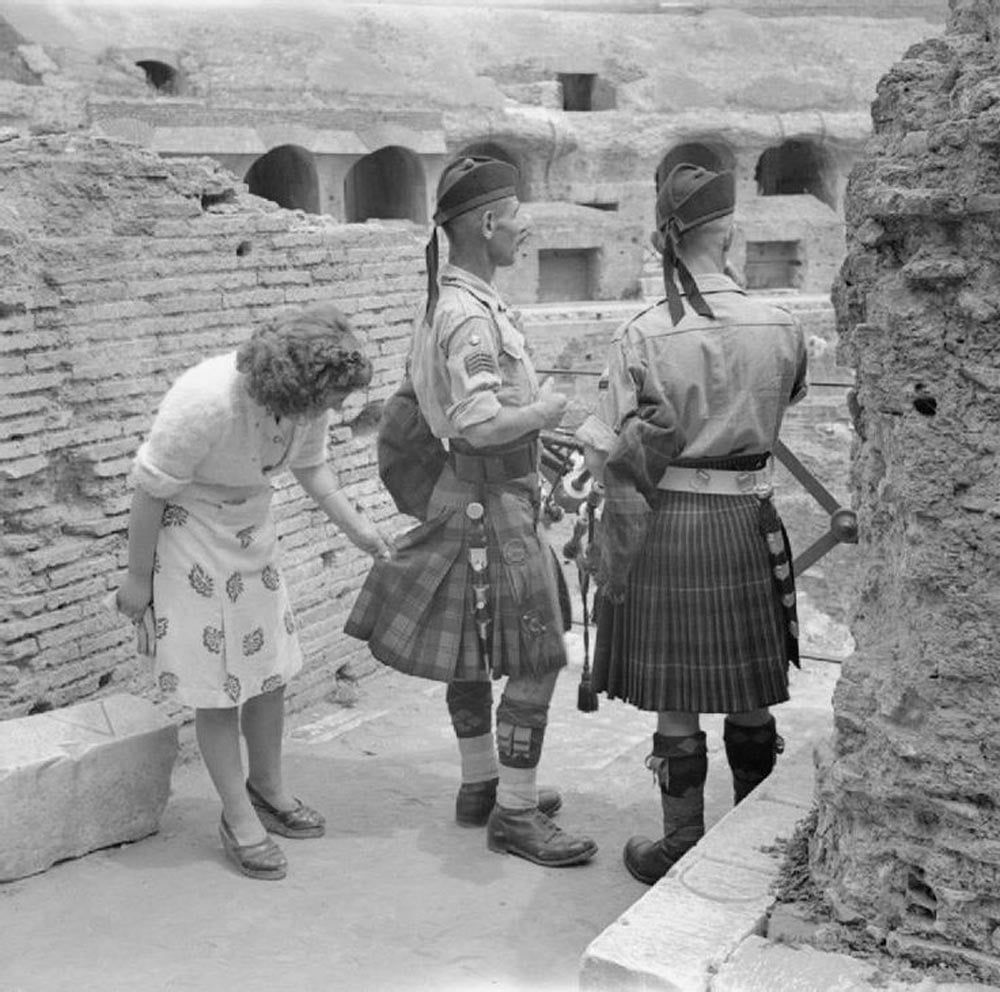 A woman playfully inspects the kilt of one of two soldiers standing next to her. The soldiers wear berets and traditional Scottish kilts, complete with sporrans. They are in a historic stone structure with multiple arched openings.
