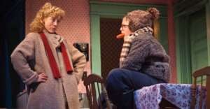 Two people in winter coats talk inside a cozy room; one stands with hands on hips, while the other sits on a table with a hat, scarf, and glasses, holding a large carrot in their mouth.