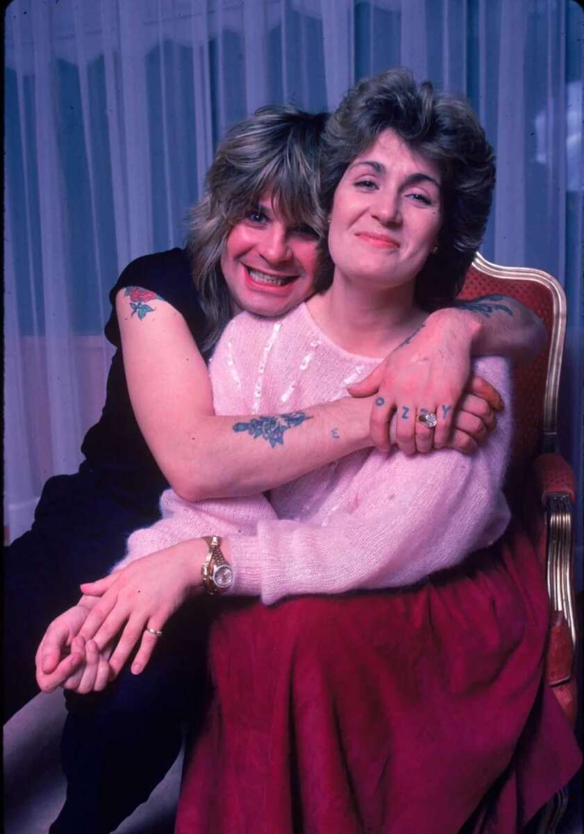 A man with long hair and tattoos hugs a smiling woman with short hair, sitting in a red chair. Both appear happy and relaxed, with a sheer curtain in the background.