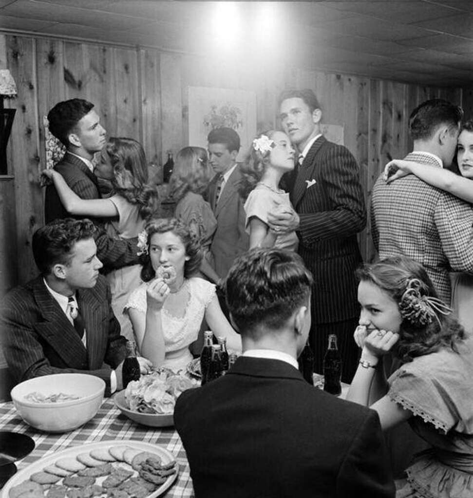 Black and white photo of a 1950s party with young adults. Three couples are dancing closely in the background. In the foreground, people are sitting at a table with snacks and soda bottles, engaging in conversation. The scene is in a wood-paneled room.