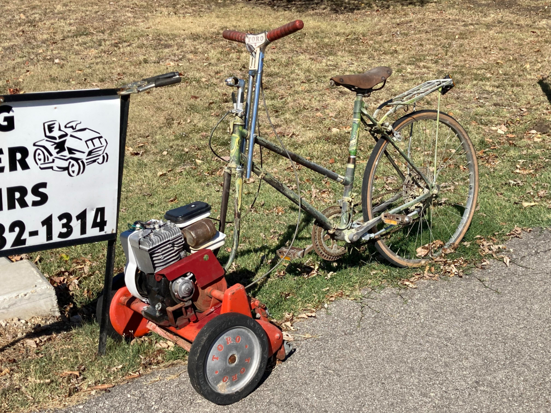 A rusty, vintage bicycle with a worn seat is missing its front wheel, which has been replaced by a red motorized machine. The bike is leaning on a roadside curb next to a sign for power equipment repairs.