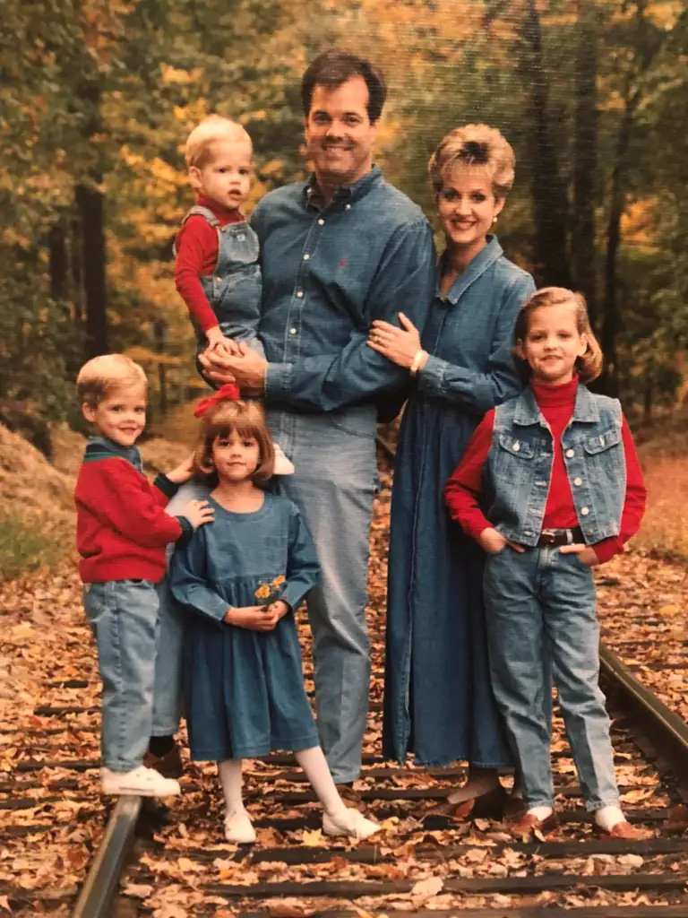 A family of six poses on railroad tracks in a forest with autumn leaves. The parents and four children all wear coordinated denim and red outfits, smiling together for the photo.