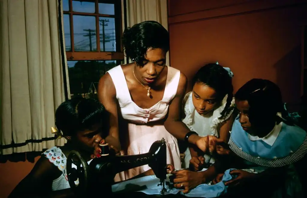 A woman teaches three girls how to use a sewing machine in a warmly lit room, all focused intently on the fabric under the machine’s needle.