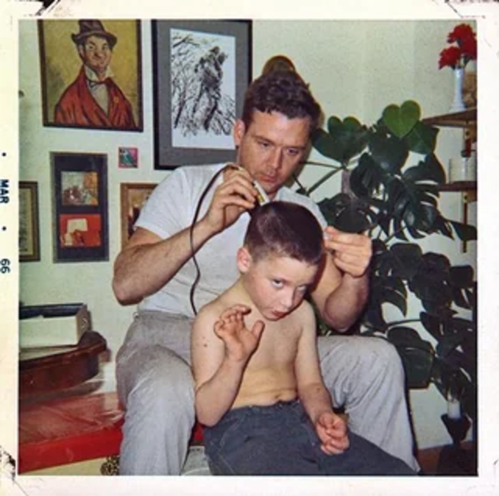 A man gives a young boy a haircut at home. The boy, shirtless, looks downward and raises one hand. Behind them are framed pictures and a leafy houseplant. The photo has a vintage look and is dated March 1966.