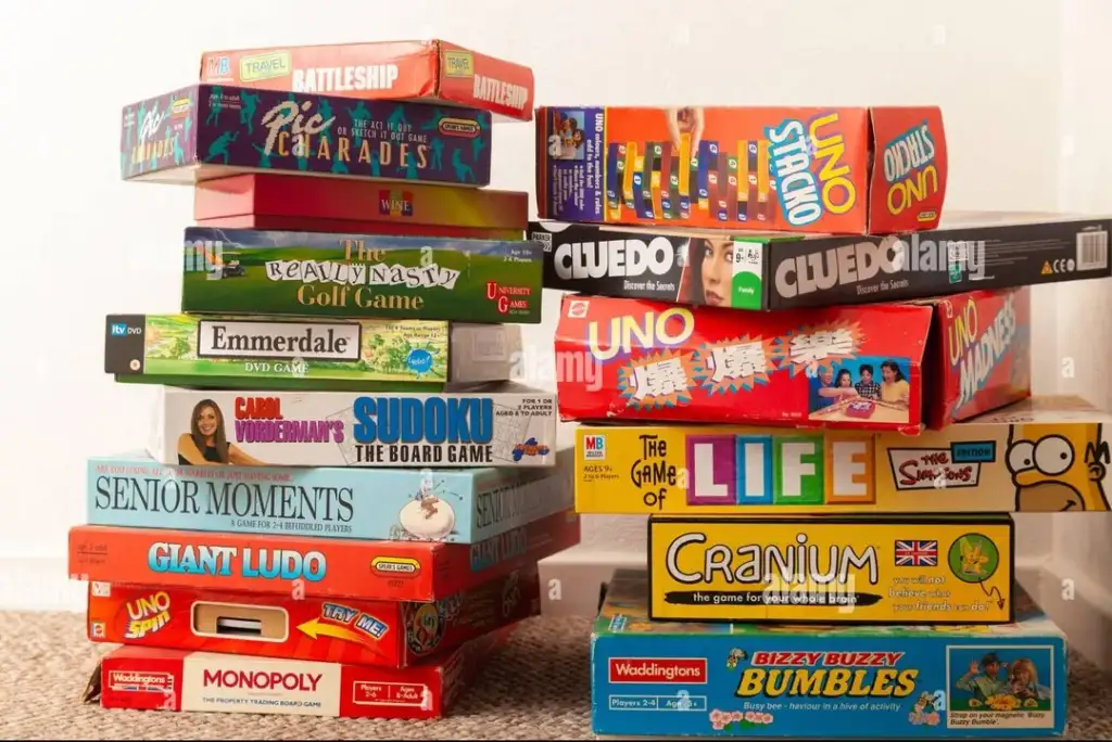 A stack of colorful board games piled on a carpeted floor, including titles like Monopoly, Cluedo, UNO, The Game of Life, Cranium, Battleship, and others.