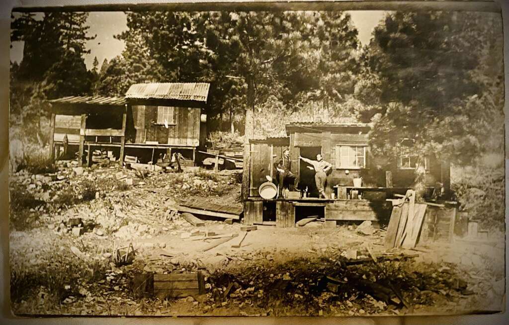 Sepia-toned image of two rustic wooden cabins in a forest setting. Three people stand on the porch of the front cabin. The area is strewn with wooden debris and has a rugged, early 20th-century frontier appearance.