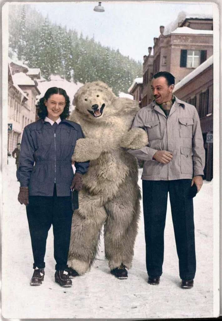 A woman and a man walking arm-in-arm with a person in a polar bear costume on a snowy village street. Flanked by snow-covered buildings, all three are smiling in winter attire.