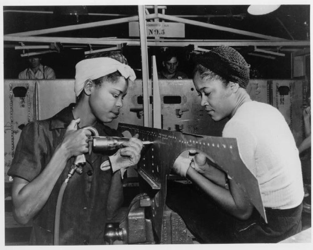 Two women work together on assembling a metal aircraft part in a factory setting. One is using a drill, while the other holds the part steady. Both are wearing protective head coverings, with focused expressions. Other workers are visible in the background.