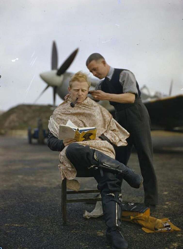 A person sits outdoors, draped in a cape, getting a haircut. They are reading a book and have a pipe in their mouth. Another person stands behind them, cutting their hair. An airplane is visible in the background.