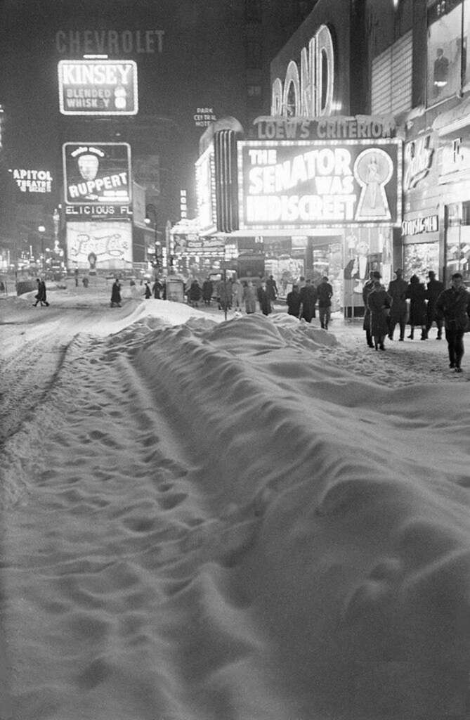 Black and white image of a snowy Times Square in New York. Snow covers the street, with people walking along the sidewalks. Bright neon signs and advertisements illuminate the scene, including a prominent theater marquee.
