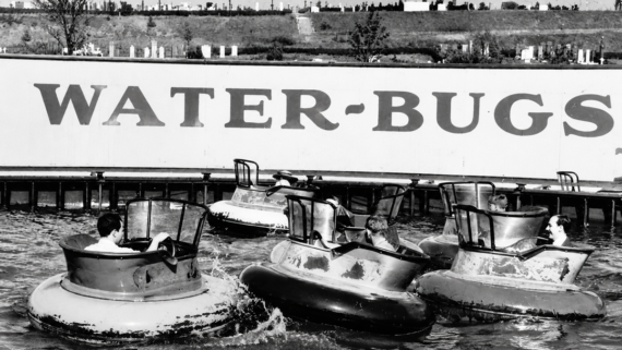 Black and white photo of people riding bumper boats with tall windshields on a water ride. A large sign in the background reads “WATER-BUGS.” Trees and fencing are visible behind the sign.