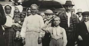 A black-and-white photo shows a group of immigrants, including men, women, and children. A boy in the front salutes, while others stand closely together, some smiling, wearing early 20th-century clothing.