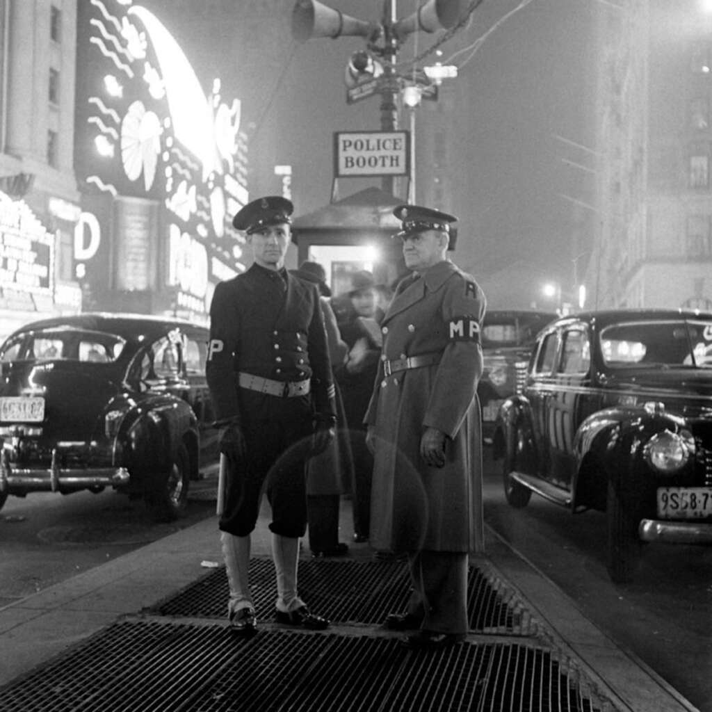 Two military police officers stand on a city street near a police booth at night. Vintage cars and bright neon signs illuminate the scene, capturing a historic, bustling urban atmosphere.
