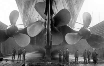 Historic black-and-white photo of the Titanic in dry dock, showing workers standing near its massive propellers and rudder beneath the ship's stern.
