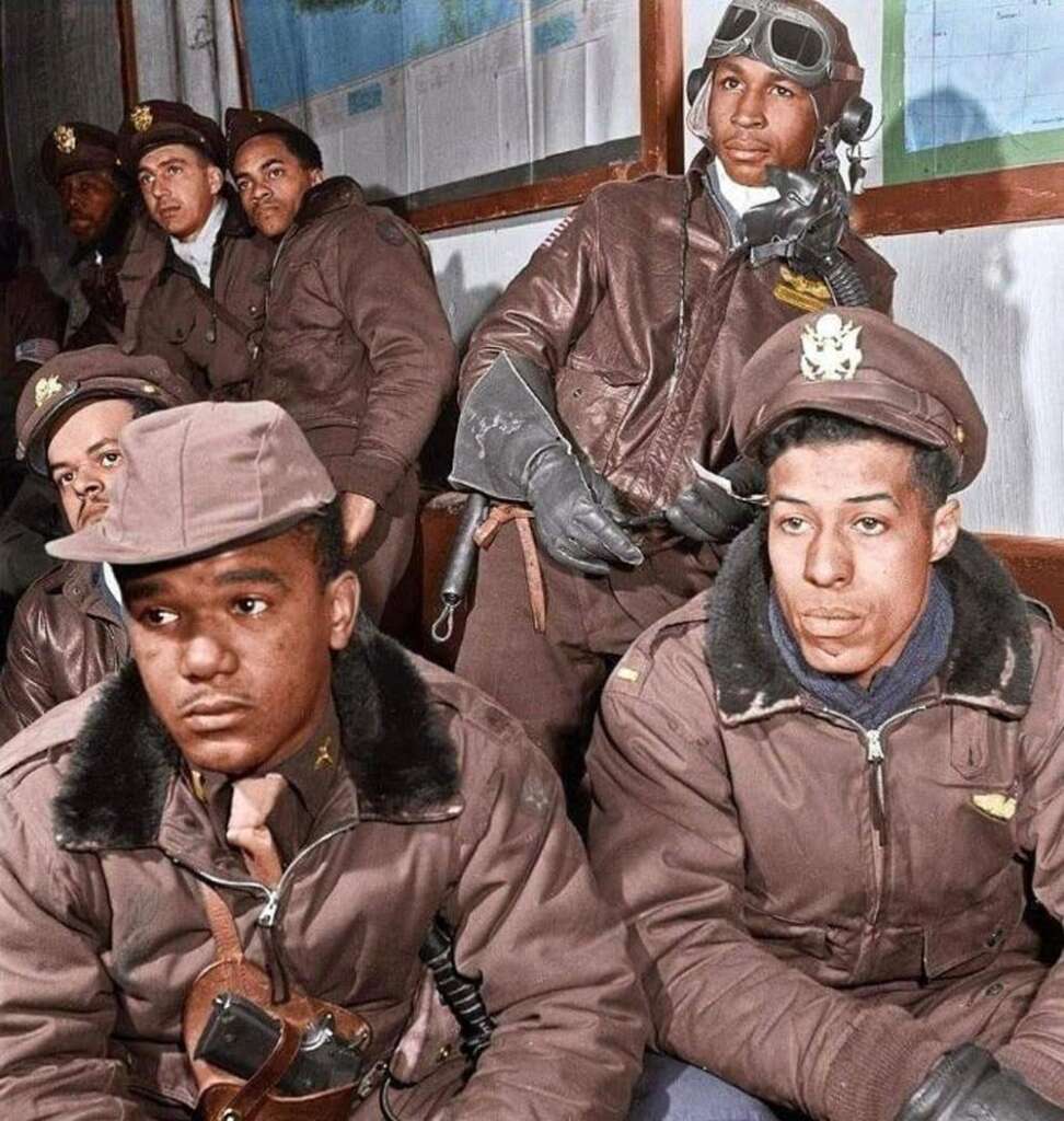 A group of military pilots in brown uniforms and hats are sitting and standing in a room. Some wear headphones and goggles. A large map is visible on the wall behind them. They appear focused and attentive.