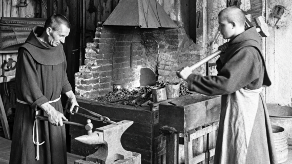 Two monks in robes work in a blacksmith shop. One holds tongs and metal on an anvil; the other prepares to strike it with a hammer. A fire burns in the forge behind them. The scene is in black and white.