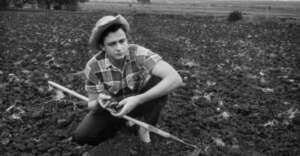 A person wearing a plaid shirt and hat crouches in a plowed field, holding soil in one hand and a hoe in the other. The background shows a vast, open landscape under a cloudy sky.