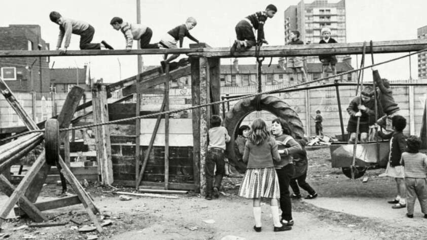 Children play on a makeshift playground in an urban environment. Some climb a wooden structure, while others gather below or engage with various equipment. Tall buildings are visible in the background. The setting is lively and dynamic.