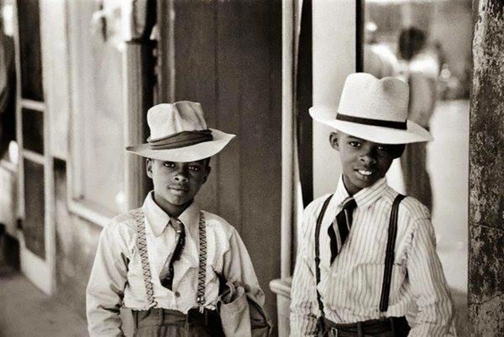 Two boys stand in front of a building, wearing wide-brimmed hats, button-up shirts, ties, and suspenders. One boy wears a dark tie and plaid suspenders, while the other has a striped shirt and white hat, both looking at the camera.
