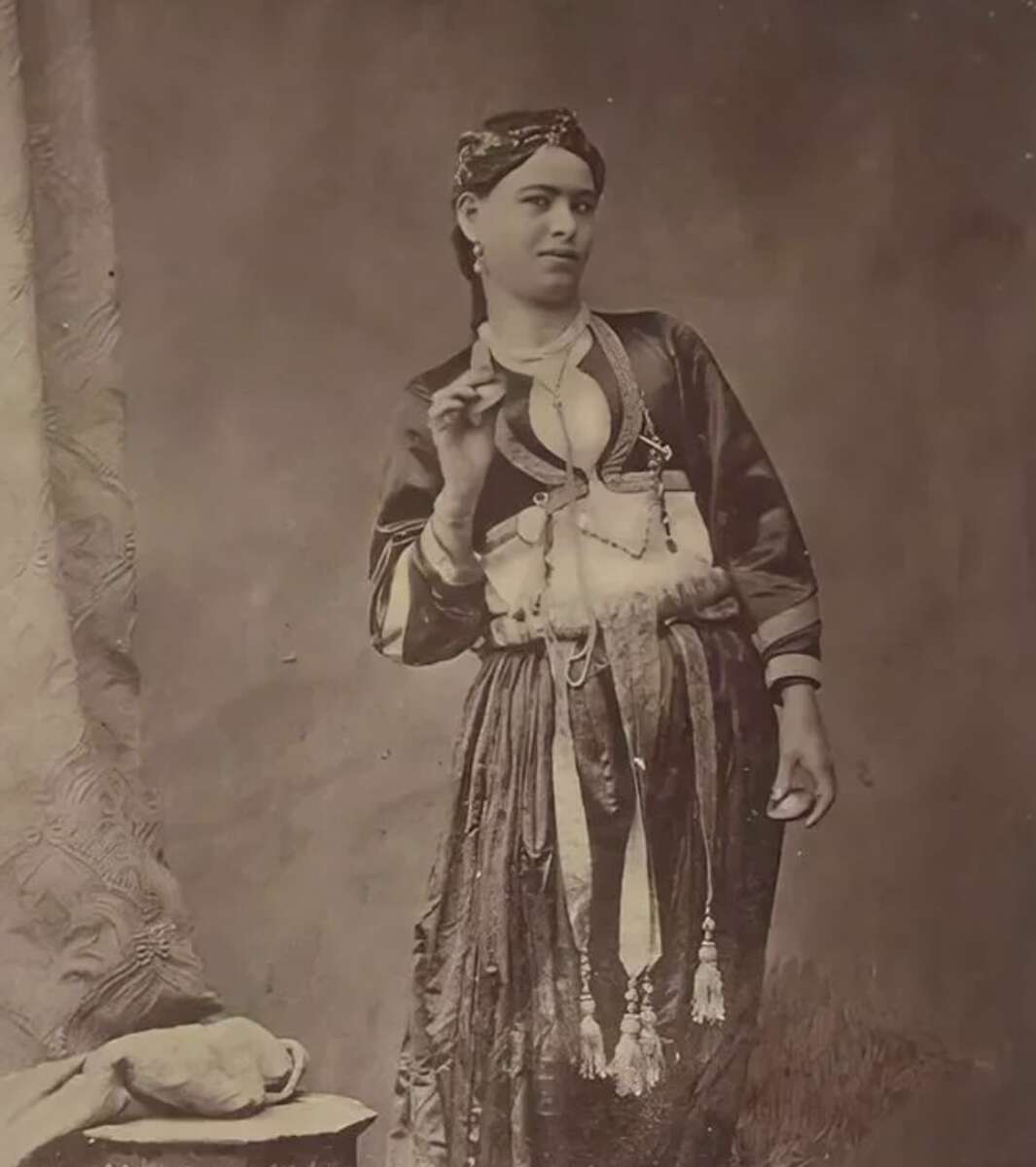 A sepia-toned vintage photograph of a person wearing traditional clothing with layered jewelry and a headscarf, standing indoors beside a small table draped with fabric.