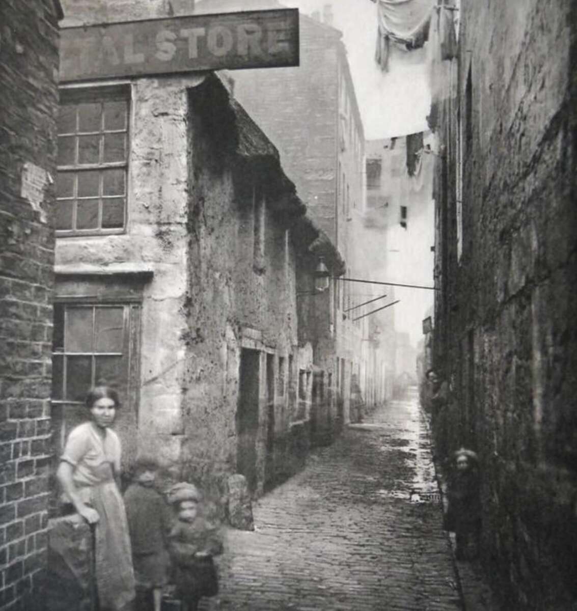 A black-and-white photo of a narrow, cobblestone alley with tall, old buildings on either side. A woman and two children stand at the entrance, and laundry hangs overhead further down the alley.