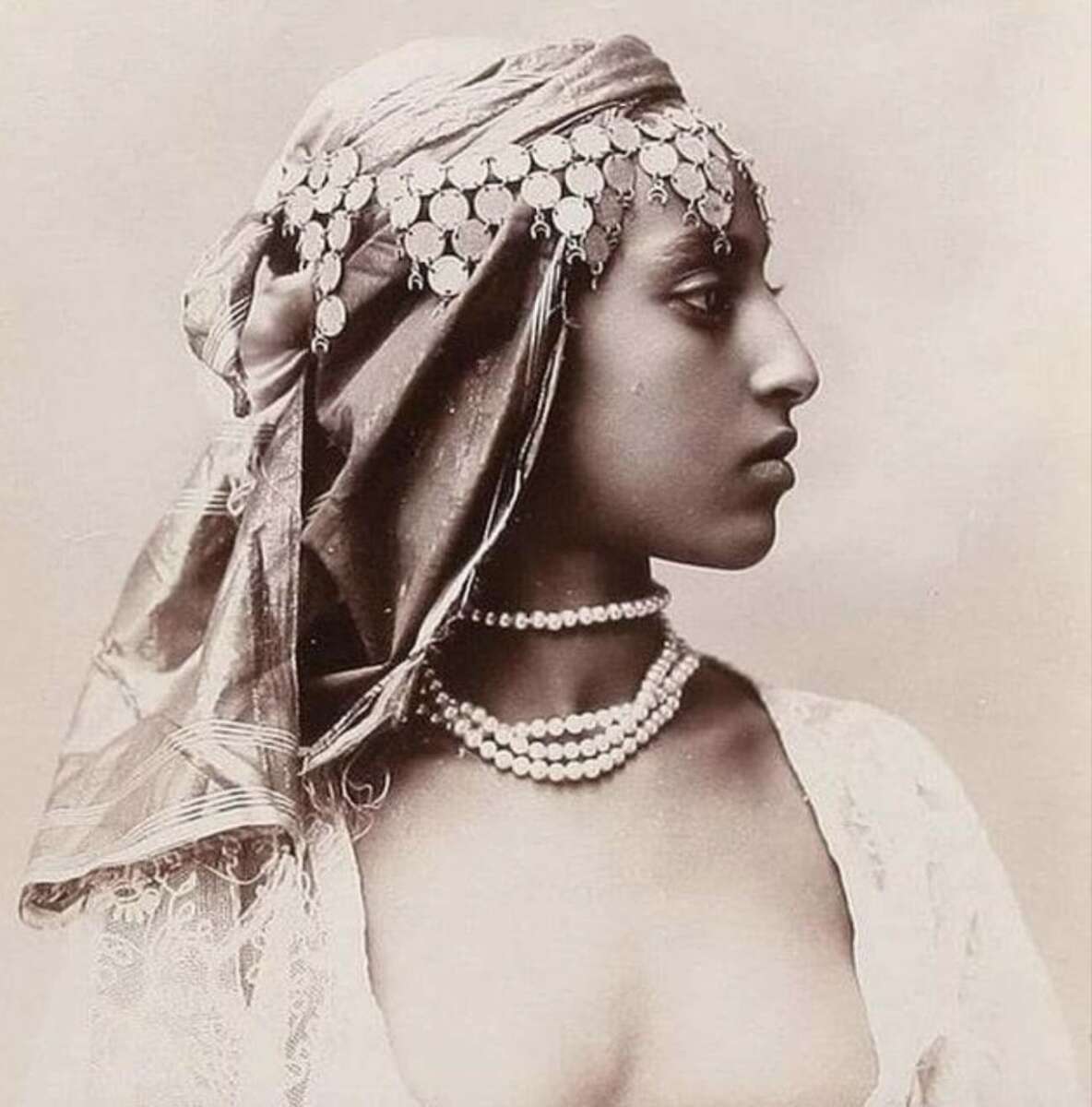 Sepia-toned portrait of a woman in profile, wearing a headscarf adorned with coins and layered pearl necklaces, with bare shoulders and a calm, dignified expression.