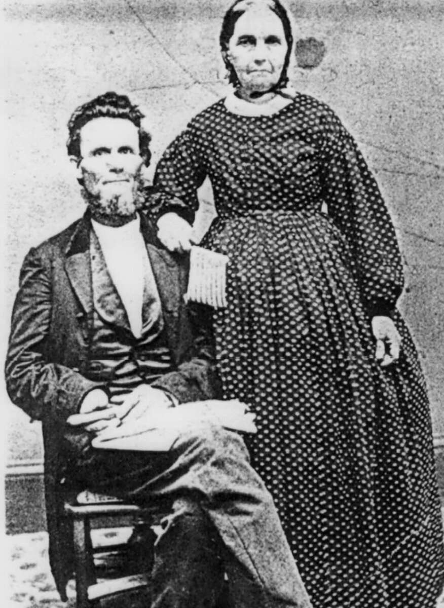 Black-and-white photo of a bearded man seated in a suit with a woman standing beside him, wearing a long, patterned dress with her hand resting on his shoulder. Both look directly at the camera.