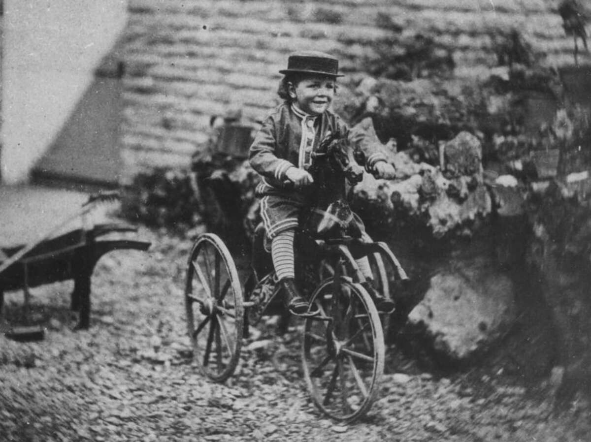 A young child wearing a hat, jacket, and striped socks smiles while riding an old-fashioned tricycle on a gravel surface, with rocks and a stone wall in the background.