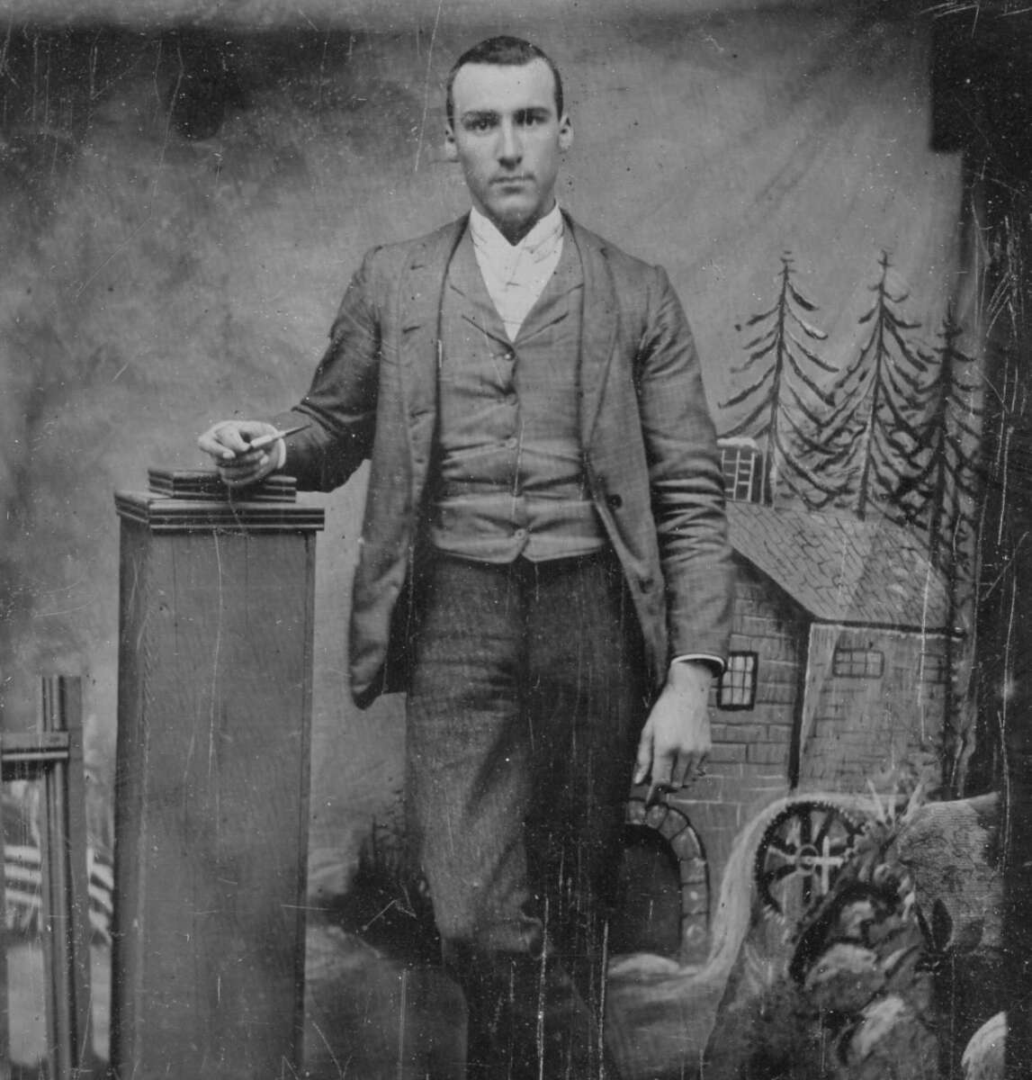 A man in a suit stands with one hand resting on a pedestal, posing for a vintage black-and-white studio portrait with a painted backdrop of a house, trees, and a waterwheel.