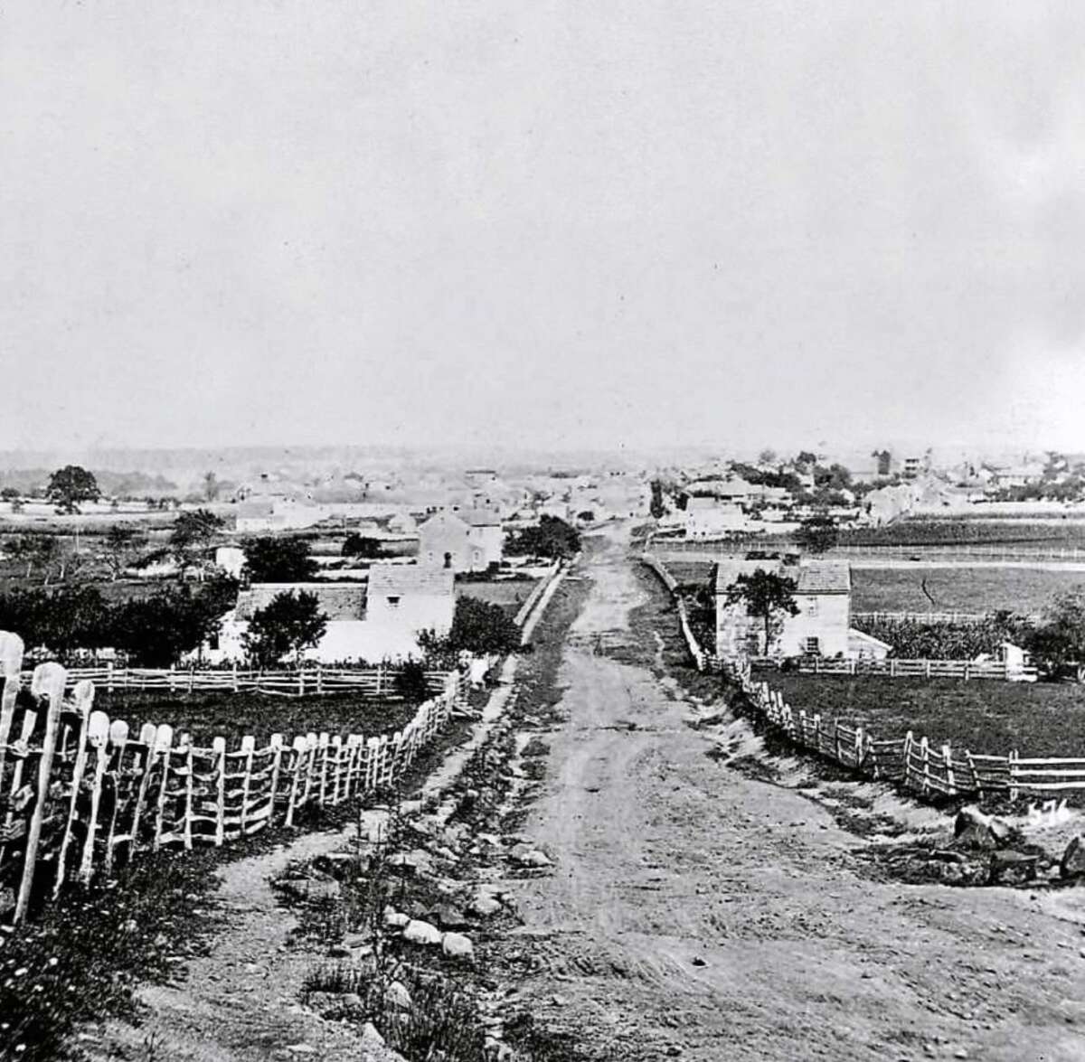 Black and white photo of a dirt road leading into a small rural town, with wooden fences, scattered houses, fields, and trees on either side, under a clear sky.