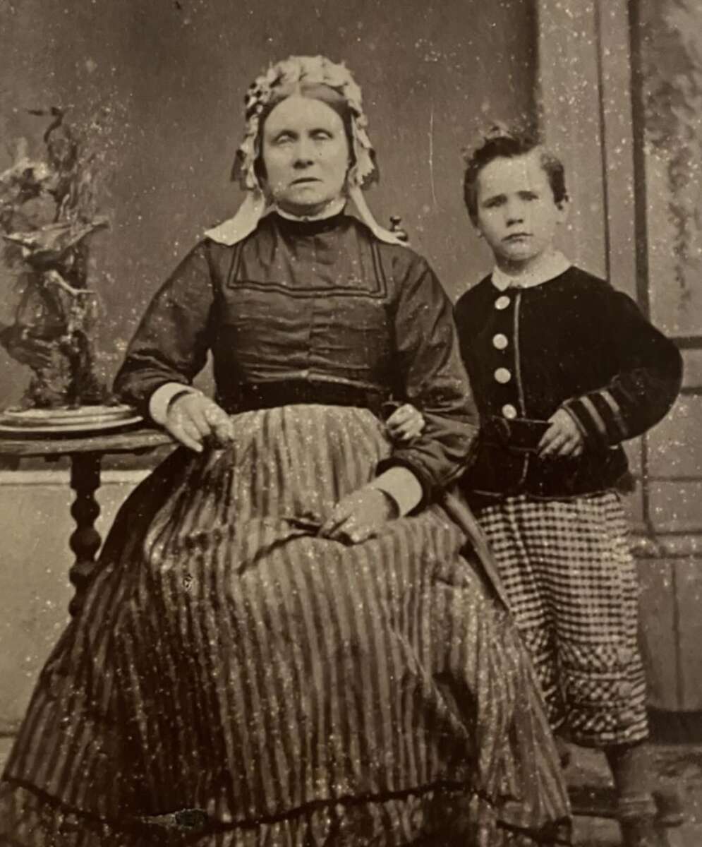 A sepia-toned vintage photograph of an older woman in a striped dress and bonnet seated next to a young boy in checkered pants and a buttoned jacket, posing indoors by a small ornate table with a decorative object.
